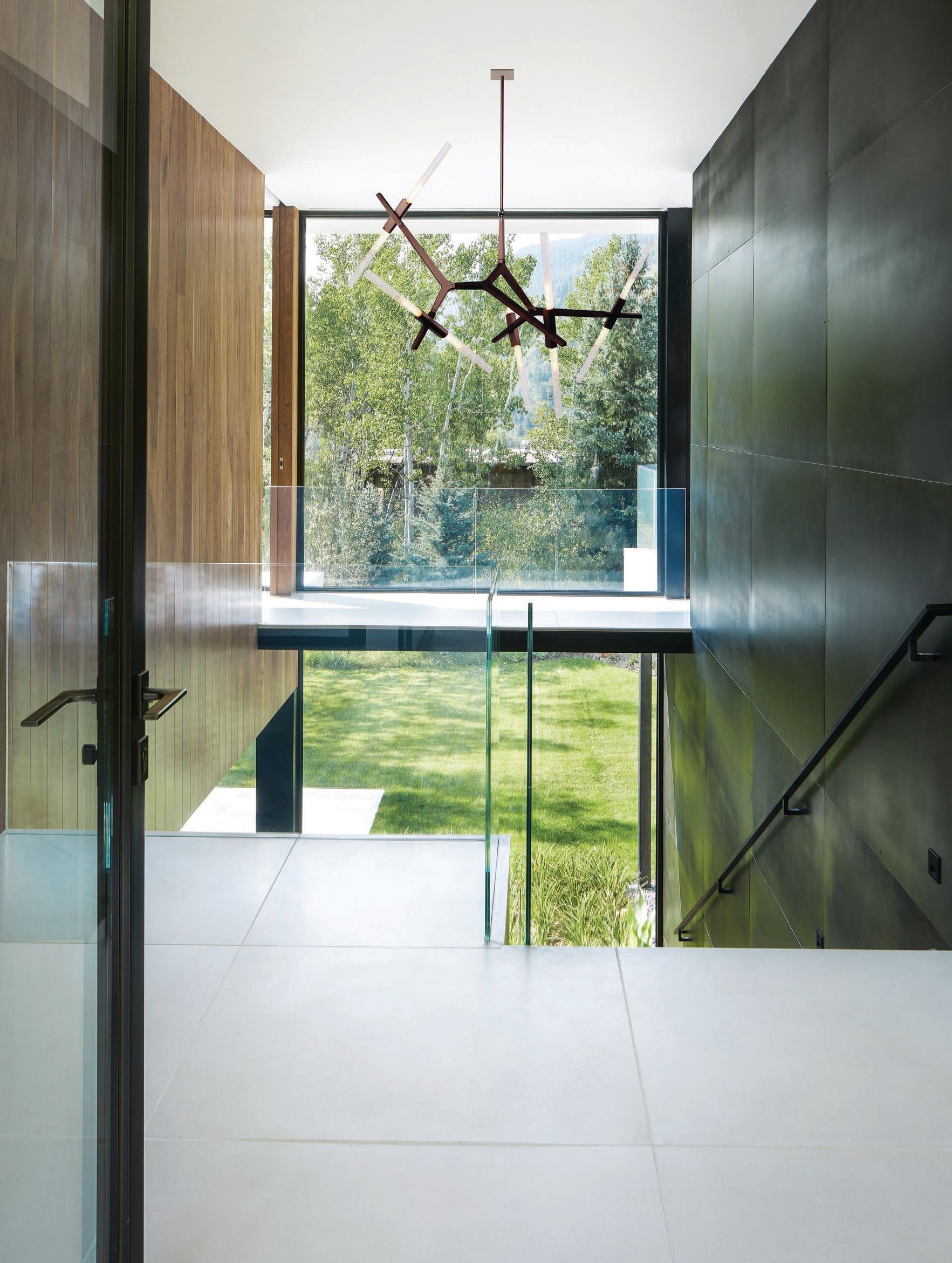 The double-height black patina steel and walnut entry introduces materials prevalent within the house and a glimpse of the lawn at the rear. The bronze Agnes 10-light chandelier by Lindsey Adelman is from Roll & Hill. PHOTOGRAPHED BY DAVID MARLOW